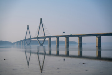 Fototapeta premium Long highway bridge spanning over a bay, with traffic driving on it. Calm water and morning haze.