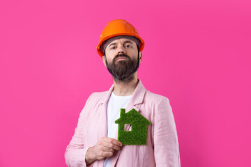 Portrait of young construction engineer wear orange hard hat, in a pink jacket standing on red studio background. A man holds a green eco house.