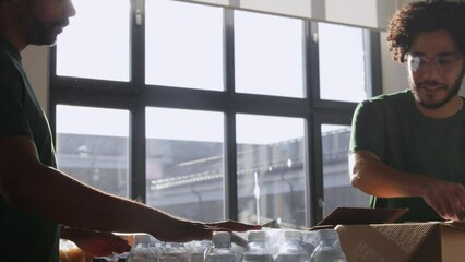 charity, donation and volunteering concept - male volunteers with clipboard packing food and drinks in boxes at distribution or refugee assistance center