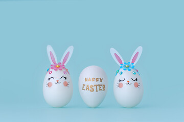 Three Easter eggs in a row on a blue background with copy space. Eggs decorated like rabbits on the sides of a white egg with a golden inscription Happy Easter. Bunny-eggs.