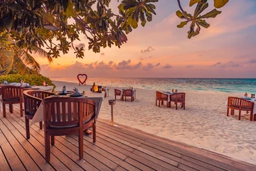 Fototapeten Outdoor restaurant at the beach. Table setting at tropical beach restaurant. Led light candles and wooden tables, chairs under beautiful sunset sky, sea view. Luxury hotel or resort restaurant © icemanphotos