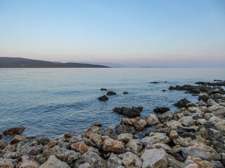 Fototapeta na wymiar Stony beach in Krk, Croatia. The Mediterranean Sea has a turquoise color. Surface of the sea is calm, there are no waves. In the back there are some mountains. Soft colors of the sunset.