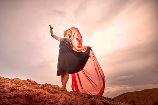 Young slim girl in black dress with red fabric dances on sand dunes against a dramatic sky before a thunderstorm. Model posing during photo shoot on nature