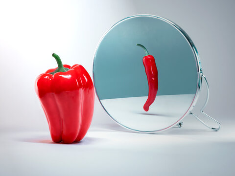 Large Red bell pepper in front of a mirror with a reflection of a thin red hot chilli pepper to illustrate body dysmorphia