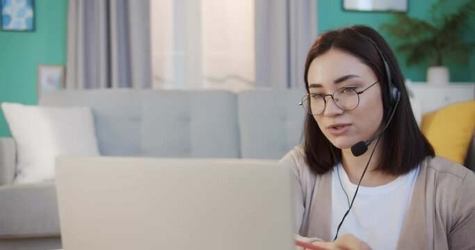 Profeshional female sales representative in headset speaking with client, making video conference call, looking at laptop, writing notes, talking on webcam. Sell online, work from home, telemarketing.