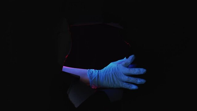 Lab Horror. Experiment Failure. Biohazard Explosion. Scientist Creepy Hand In Glove Breaking Through Black Ripped Wall With Neon Color Light Isolated On Dark Empty Space.