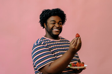 young happy african american man eating strawberries in the studio over pink background
