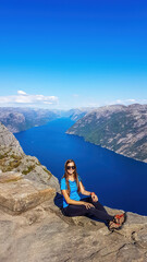 Fototapeta premium A girl wearing blue T-shirt sitting at the edge of a steep cliff of Preikestolen, with a view on Lysefjorden. Fjord goes far inland. Girl enjoys the view, feeling free and happy. Great accomplishment
