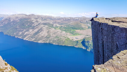 A man wearing blue shirt sits at the edge of a steep cliff of Preikestolen with his legs hanging down. A view on Lysefjorden. Fjord goes far inland. Man enjoys the view, feels free and happy.