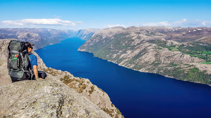 A man wearing blue shirt, full cap and a huge hiking backpack sits at the edge of a steep cliff of Preikestolen. A view on Lysefjorden. Fjord goes far inland. Man enjoys the view, feels free and happy