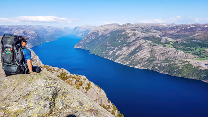 Fototapeta premium A man wearing blue shirt, full cap and a huge hiking backpack sits at the edge of a steep cliff of Preikestolen. A view on Lysefjorden. Fjord goes far inland. Man enjoys the view, feels free and happy