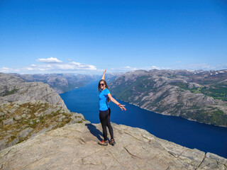 A girl wearing blue T-shirt standing at the edge of a steep cliff of Preikestolen, with a view on Lysefjorden. Fjord goes far inland. Girl enjoys the view, feeling free and happy. Great accomplishment