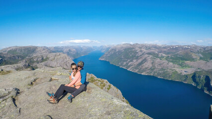 A couple sitting at the edge of a steep cliff of Preikestolen, with a view on Lysefjorden. Fjord goes far inland. The couple enjoys the view, feeling free and happy. Great accomplishment