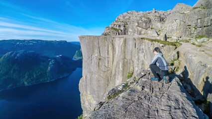 A young man in a hiking outfit squats at the edge of a steep cliff with the view on Lysefjorden and...