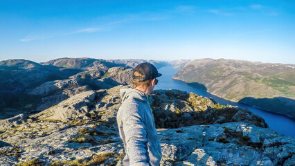 Naklejka premium A young man in a hiking outfit walks along the edge of a steep cliff , with a selfie stick. He walks on a barren rocky cliffs. In the back is Lysefjorden, Norway. Bright and sunny day.