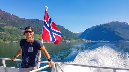 A young man leaning on a rail of a ship with a Norwegian flag waving behind him, Songefjorden, Norway. The motor of the ship makes the water wavy and foamy. Lush green mountains surrounding the fjord. - obrazy, fototapety, plakaty