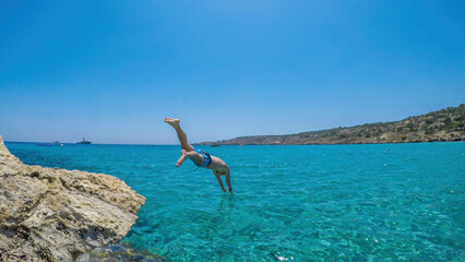 A young athletic-build man jumping into the sea in Cape Greco, Cyprus. Water is crystal clear, shimmering with many shades of blue. Jump of joy. Man is wearing blue swimsuit.