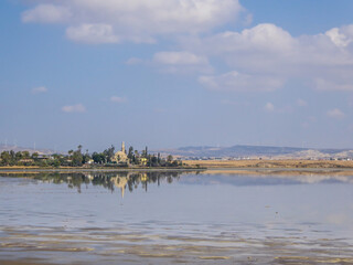 Hala Sultan Tekke seen from a distance. The mosque is surrounded by lush setting - palm trees and smaller bushes. Clear reflection of the mosque in the calm surface of a Larnaca Salt Lake.
