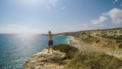 Fototapeta premium A young man standing at the steep cliff and spreading his arms wide in a gesture of freedom in Paphos, Cyprus. Blue sea spreads in front of him. Barren slopes of the cliff. Endless horizon line.