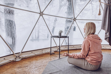 A woman sits in a geo dome glamping tent and meditates, does pranayamas, looks at the winter snow-covered nature