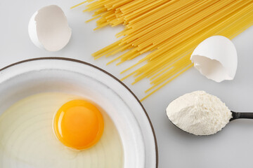 White broken egg with a yolk in a white plate with a spoonful of flour and spaghetti or pasta. Cracked egg, eggshells with yolk.  Shallow depth of field