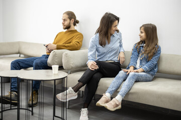 Young people sitting on the couch at the waiting room of the clinic. Man and woman with her daughter waiting for a doctor's appointment at reception