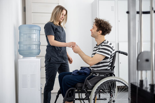 Nurse helping guy in wheelchair to fill water from cooler at rehabilitation center. Concept of support and assistance to people with disabilities