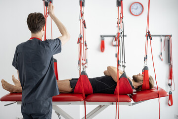 Rehabilitation specialist preparing for an active treatment on suspension straps with a male...