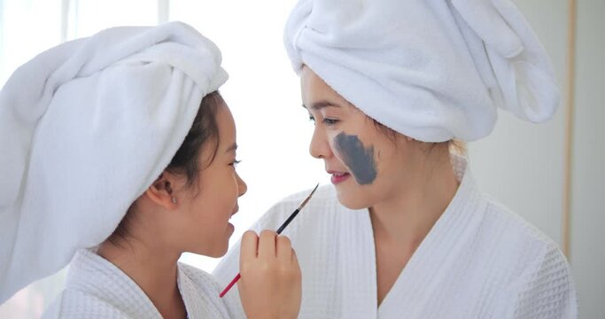 Daughter applied grey mask cream on her mom face having fun in the bedroom after shower. Family beauty treatment.