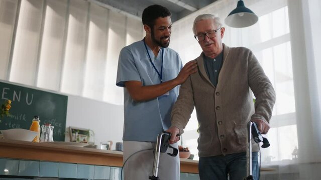 Caregiver helping senior man to walk with walker indoors in retirement nursing hme.