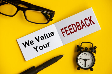 Text on notepad with reading glasses, pen and alarm clock on yellow background - We value your feedback. Conceptual