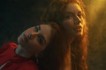 Two beautiful redhead freckled women with long natural curly hair posing in smoke, darkness and warm light. Art studio portrait made through the wet glass. Film grain effect
