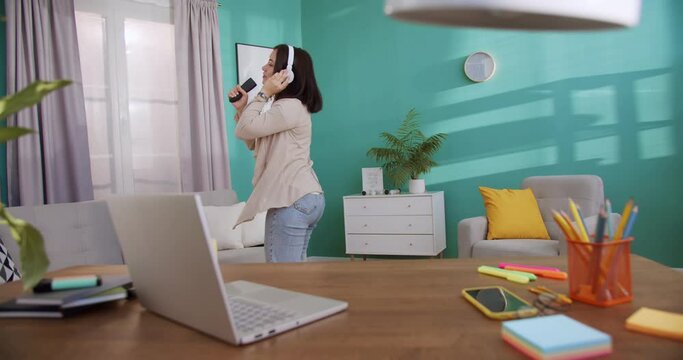 Funny playful brunette woman in headphones listening music, dancing and singing at home having fun, celebrating success, enjoying karaoke music entertainment in home office.