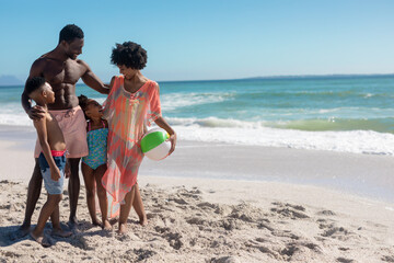 Happy african american parents and children standing together with ball at beach on sunny day