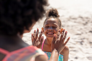 Cheerful african american girl smiling with eyes closed while mother applying suntan lotion on face
