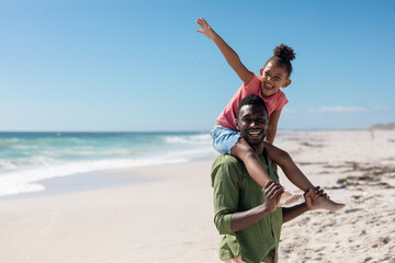 Cheerful african american girl sitting on father's shoulders enjoying summer holiday at beach