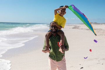 Portrait of african american man carrying son with kite on shoulders at beach during sunny day