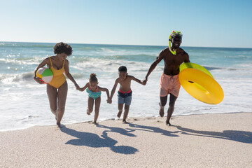 Happy african american family holding hands while running together at beach against clear sky