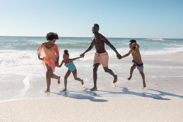 African american parents and children holding hands while running together at beach on sunny day - Powered by Adobe