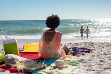 Rear view of african american mother sitting while looking at children playing at beach