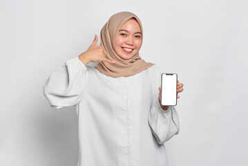 Smiling young Asian Muslim woman showing mobile phone blank screen recommending App and gesturing call me isolated over white background