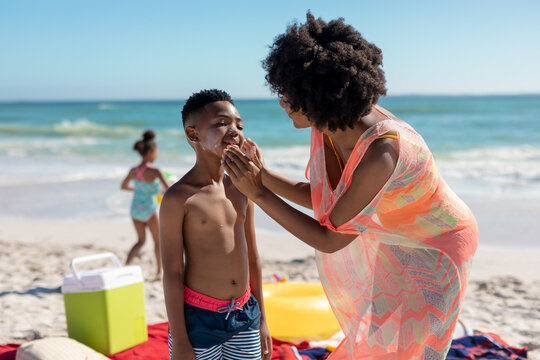 African american woman applying suntan lotion on son's cheeks at beach during sunny day