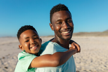 Side view of portrait of smiling african american father piggybacking son at beach on sunny day