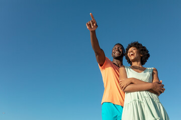 Low angle view of happy african american man pointing with girlfriend against clear blue sky