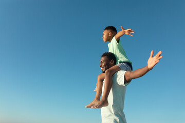 Happy african american boy on father's shoulders by copy space against clear blue sky