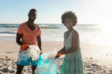 Portrait of smiling african american couple cleaning beach from plastic waste on sunny day - Powered by Adobe