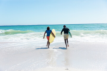 African american senior couple carrying surfboards running towards waves at beach with copy space