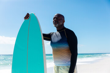 Retired senior african american man holding surfboard standing at beach against blue sky - Powered by Adobe