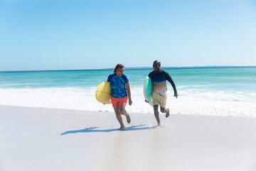 African american senior couple carrying surfboards running at beach against blue sky with copy space