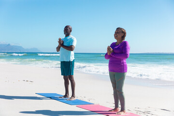 Fit african american senior couple practicing yoga at beach against blue sky on sunny day